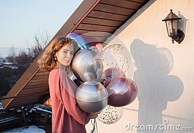 Happy birthday teenage girl in pink dress holding a bunch of colorful balloons, stands near house on a sunny day Stock Photo