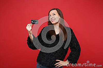 Portrait of thinkful young woman in black fur sweater looking up, holding credit bank card isolated on bright red Stock Photo