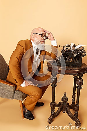 Portrait of thoughtful, bearded, mature, bald man in elegant, classical suit sitting near typewriter against studio Stock Photo