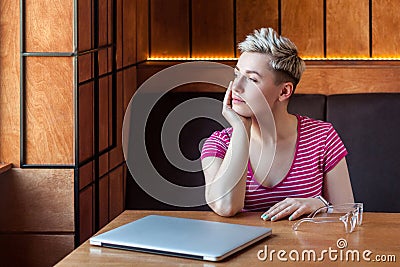 Portrait of thoughtful beautiful young bussineswoman with blonde short hair in pink t-shirt is sitting in cafe, finishing work and Stock Photo