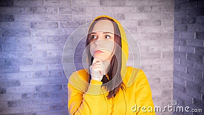 Portrait of thoughtful young woman in hood on gray brick background. Close up of cute brunette propping up her chin in Stock Photo