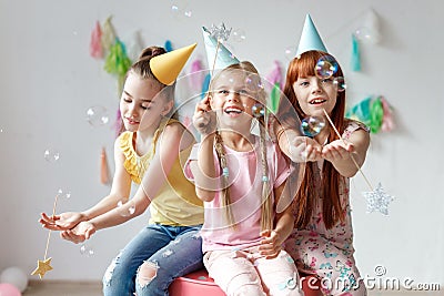 Portrait of three beautiful girls wear festive caps, play with bubbles, sit together on chair, celebrate birthday, being Stock Photo