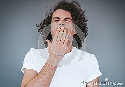 Portrait of tired or bored Caucasian young male model with curly hair covering mouth while yawning, feeling exhausted after Stock Photo