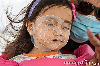 A portrait of toddler girl closing eyes feeling the wind while father in the background wearing face mask on the boat Stock Photo