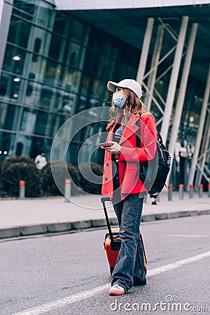 Portrait of a traveler woman in a mask walking with an orange suitcase near an airport. Young fashionable woman in a Stock Photo