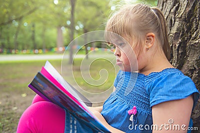 A portrait of trisomie 21 child girl outside having fun on a park reading book Stock Photo