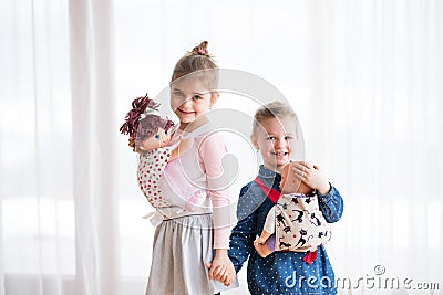 A portrait of two small girls standing and carrying dolls in baby carriers indoors. Stock Photo