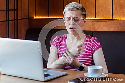 Portrait of unhappy young bussinesswoman with short blonde hair in pink t-shirt and eyeglasses sitting in cafe and holding arm Stock Photo