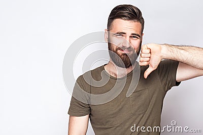 Portrait of unsatisfied bearded man with thumbs down and dark green t shirt against light gray background. Stock Photo