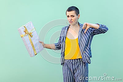 Portrait of unsatisfied with short hair young woman in striped suit standing, showing dislike gesture to her present box, looking Stock Photo