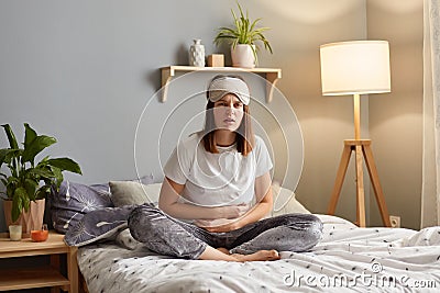 Portrait of upset Caucasian woman suffering from strong abdominal pain, touching her tummy, sitting on bed, feeling acute Stock Photo