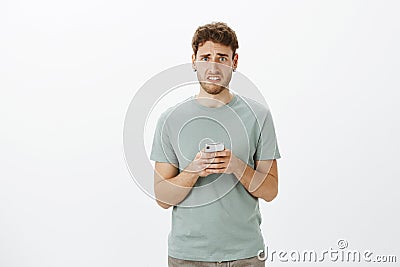 Portrait of upset gloomy cute guy with fair hair and beard, frowning and grimacing, holding smartphone, realising Stock Photo