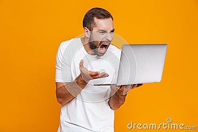 Portrait of uptight man 30s in white t-shirt screaming and holding silver laptop, isolated over yellow background Stock Photo