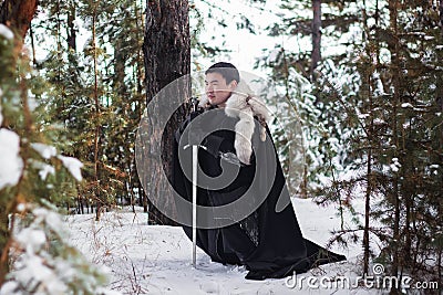 Portrait of a warrior in black quilted clothes, leather gloves, in a cloak with a fur collar. He sits on his knee and prays Stock Photo