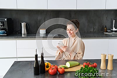 Portrait of woman in bathrobe sitting in kitchen with smartphone, cooking dinner, watching recipe on social media, video Stock Photo