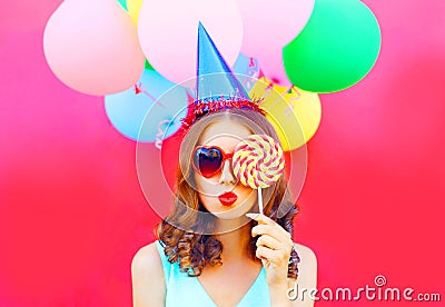 Portrait woman in a birthday cap is blowing lips is closes her eye with lollipop on stick over an air colorful balloons Stock Photo
