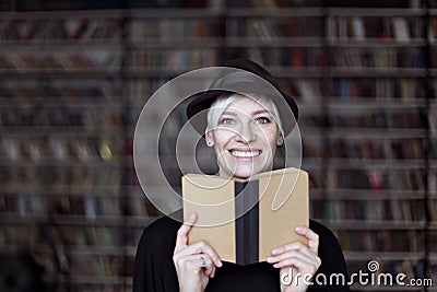 Portrait of woman in black hat with opened book smiling in a library, blonde hair. Hipster student girl Stock Photo