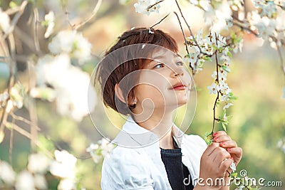 Portrait of 7 years old cute stylish short haired smiling girl in white shirt sniffing flower in blooming garden in spring. Short Stock Photo