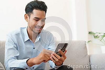 Portrait of young asian man handsome wearing blue shirt is reading using smartphone and sitting on sofa in living room at home Stock Photo