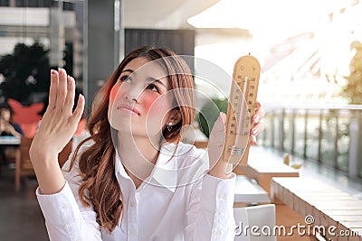 Portrait of young Asian woman holding thermometer and feeling so hot with high temperature on the desk against sunshine effect bac Stock Photo