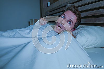 Portrait of young attractive scared man in fear and panic suffering horror nightmare waking up suddenly at night lying on bed in d Stock Photo