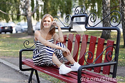 Portrait of a young attractive smiling girl sitting on a park bench. Blurred background. Stock Photo