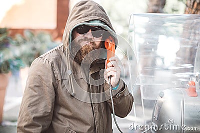 Portrait of young bearded man using public phone wearing hoodie jacket and black sunglasse Stock Photo