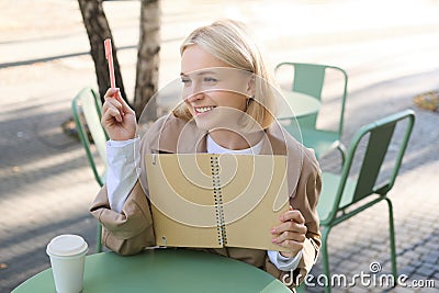 Portrait of young beautiful blond woman, artsy girl in coffee shop, holding notebook and pen, writing in her journal Stock Photo