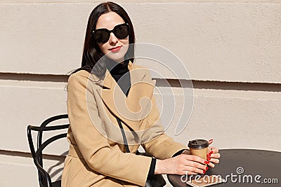 Portrait of young beautiful brunette woman in stylish sunglasses and beige fashionable coat sits at table in outdoor cafe. Stock Photo