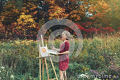 Middle age white Caucasian woman artist drawing in open plein air outside with watercolor on paper at easel. Stock Photo