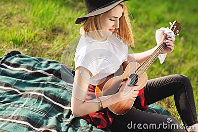 Portrait of a young beautiful woman in a black hat. Girl sitting on the grass and playing guitar Stock Photo