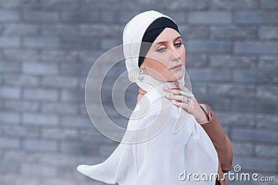 Portrait of a young blue-eyed woman in a hijab against a gray brick wall. A Muslim woman looks at the camera turning Stock Photo