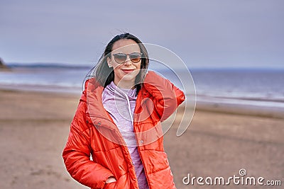 Portrait of a young brunette woman in sunglasses and a red jacket. Woman on a walk along the sandy shore of a large river on a spr Stock Photo