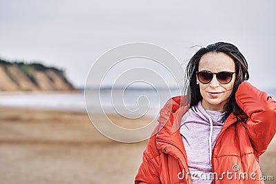 Portrait of a young brunette woman in sunglasses and a red jacket. Woman on a walk along the sandy shore of a large river on a spr Stock Photo
