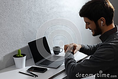 Portrait of young businessman verifies time on smartwatch, using wireless earphones. Laptop, coffee mug, cactus and smartphones. Stock Photo