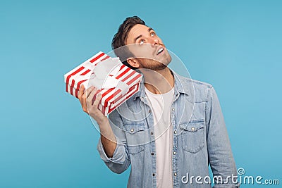 Portrait of young curious man in denim shirt holding gift box near ear and listening, guessing present Stock Photo