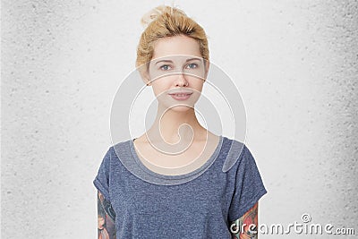 Portrait of young cute girl with blonde hair, pierced nose and tattoos, wearing casual blue t-shirt smiling and watching directly Stock Photo