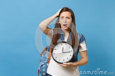 Portrait of young dissatisfied woman student with backpack clinging to head, holding alarm clock isolated on blue Stock Photo