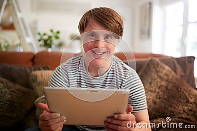 Portrait Of Young Downs Syndrome Man Sitting On Sofa Using Digital Tablet At Home Stock Photo