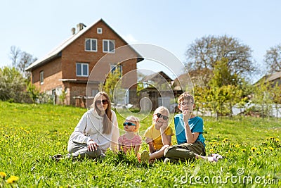 Portrait of young family sitting in the grass against two-story house. Mom or nanny sitting with children in meadow surrounded by Stock Photo