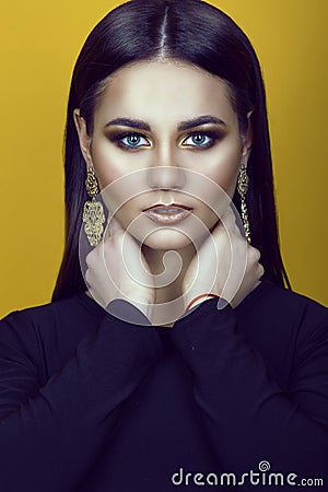 Portrait of young gorgeous blue-eyed dark-haired model with professional make up in golden colors wearing black top and earrings Stock Photo