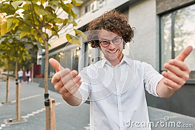 Portrait of young handsome businessman standing outdoors looking and waving in the camera. Smiling friendly man with curly hair Stock Photo