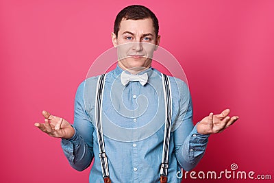 Portrait of young handsome man with astonished facial expression, shruggs shoulders with hesitation, dressed in formal shirt in Stock Photo