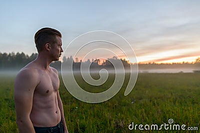 Profile view of young handsome muscular man shirtless against grass field with fog in the break of dawn Stock Photo