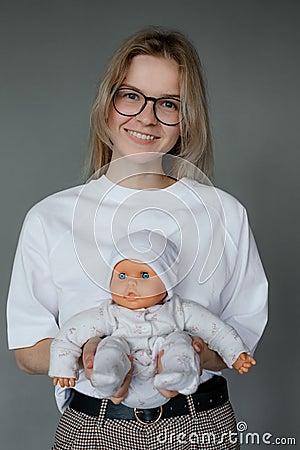 Portrait of young happy beauteous woman holding baby doll in white clothes with two hands on grey background. Studio. Stock Photo