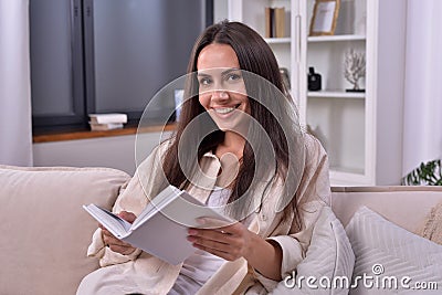 Happy young woman smiles at camera, holding white book with blank cover. Lounging on sofa in home interior. Embracing reading conc Stock Photo