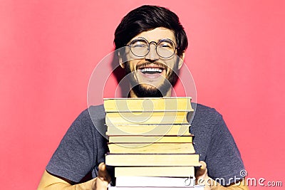 Portrait of young laughing man, holding bunch of books, wearing round eyeglasses on background of coral pink color. Stock Photo