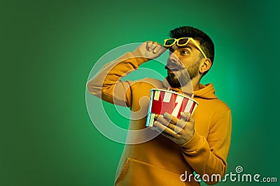 Portrait of young man in glasses with popcorn box isolated over green background in neon light. Shocking movie scene Stock Photo