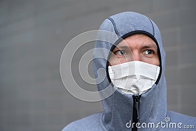 Portrait of young man in zipped gray hoodie and home made cotton face mouth virus mask, only eyes visible, gray wall in background Stock Photo