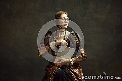 Portrait of young, pretty girl, royal person in vintage dress and modern glasses posing with laptop against dark green Stock Photo
